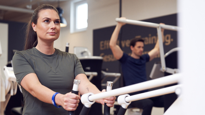 OptiMum Kundin und Kunde trainieren an Geräten OptiMum Kundin im Vordergrund trainiert an einem Gerät und schaut fokussiert, im Hintergrund trainiert ein Kunde an einem egym Gerät
